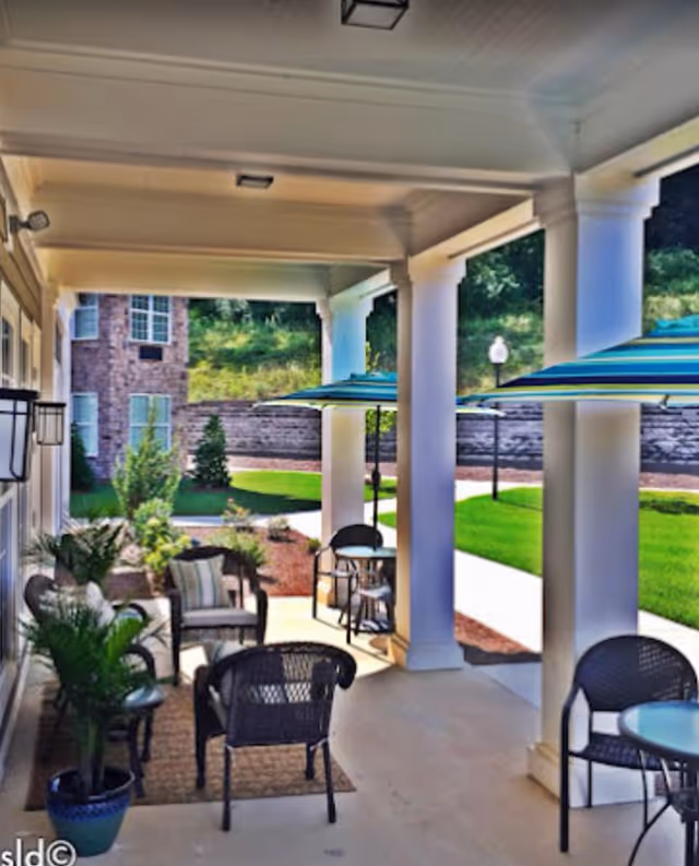 Covered outdoor patio with columns, wicker chairs and tables, potted plants, and striped umbrellas overlooking a grassy courtyard.