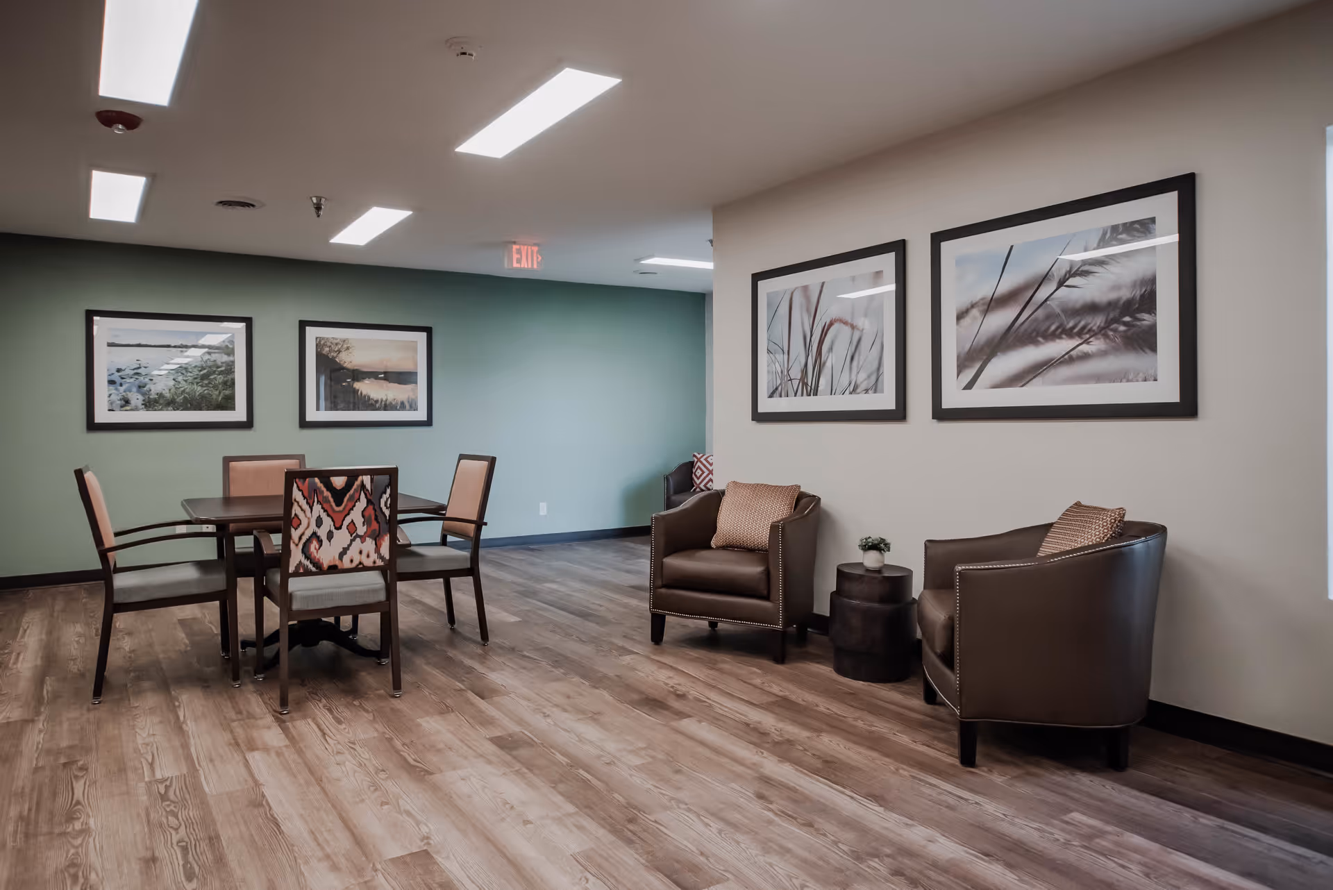 A well-lit interior room with wooden flooring featuring a small dining table with four chairs, two leather armchairs with cushions, a small round side table with a plant, and framed nature-themed artwork on the walls.