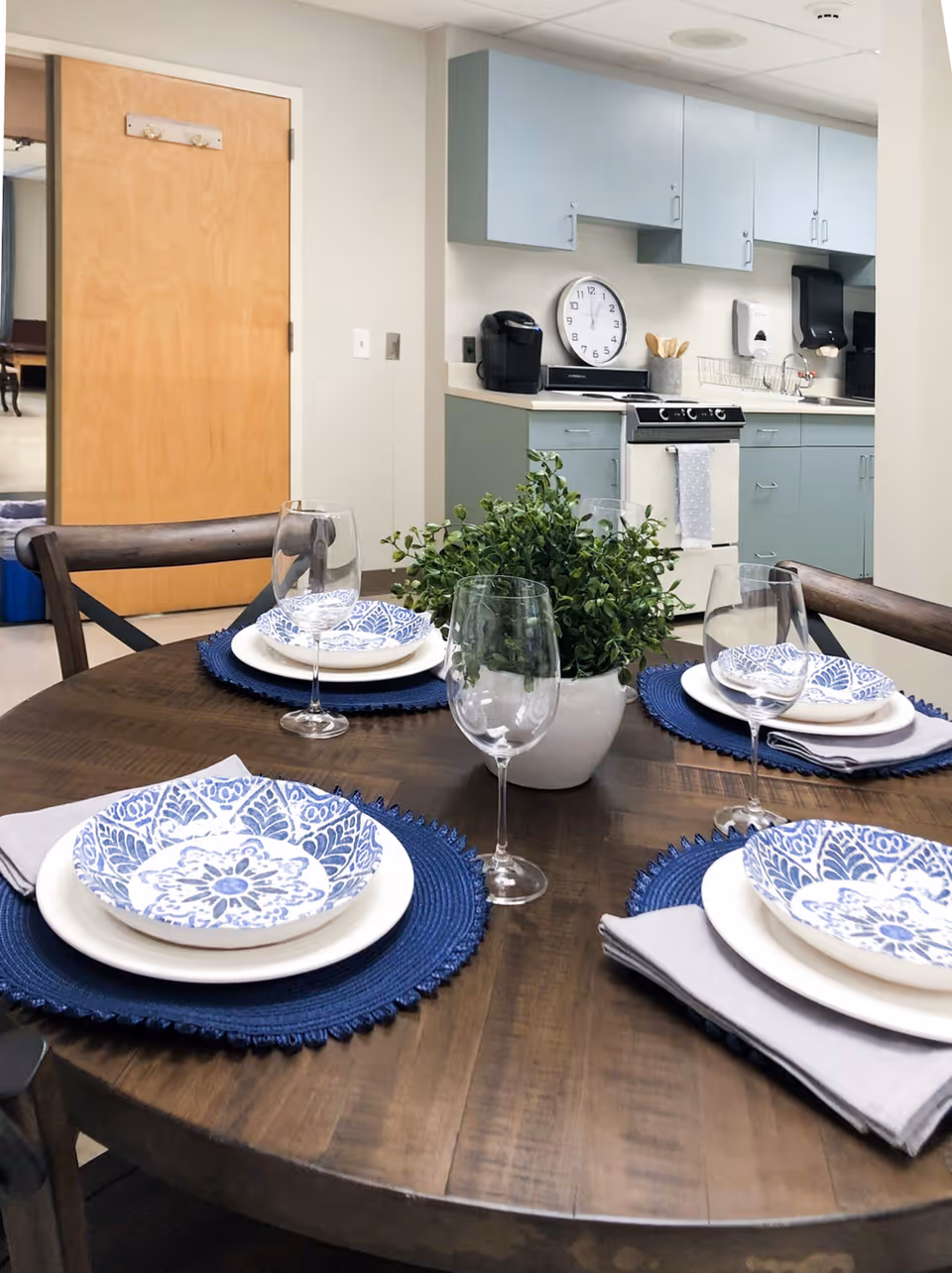 A dining table set for four with blue and white patterned plates, wine glasses, and gray napkins on blue placemats. A potted green plant is in the center of the wooden table. In the background, there is a kitchen area with light blue cabinets, a stove, a coffee maker, and a wall clock.