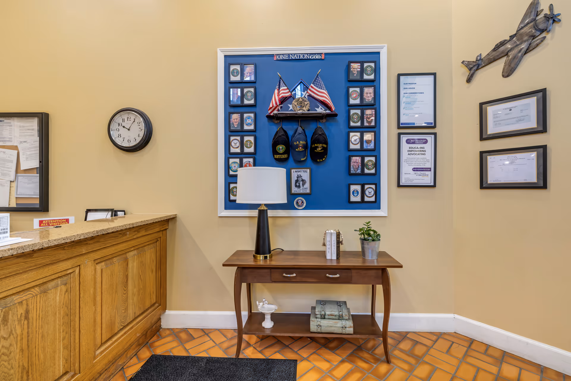 Reception area interior with a wooden front desk, a small table with a lamp and plant, and a blue display of military hats and American flags on the wall.