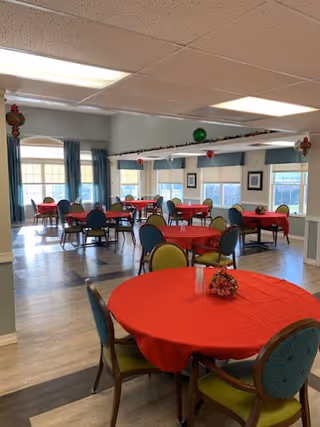A dining room with multiple round tables covered with red tablecloths and decorated with small floral centerpieces. The room has large windows with blue curtains, light-colored walls, and a ceiling with fluorescent lights. Chairs with green and blue upholstery surround the tables.