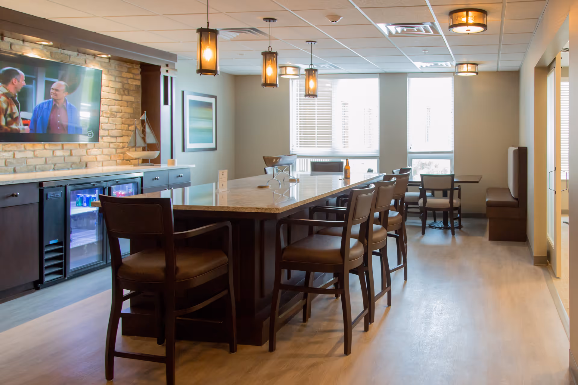 A modern communal dining area with a large rectangular table surrounded by chairs. The room features pendant lights hanging from the ceiling, a brick accent wall with a mounted flat-screen TV showing a scene from a TV show, and a countertop with built-in mini refrigerators. There is a small table with chairs and a bench seat near the windows with blinds, allowing natural light to fill the space.
