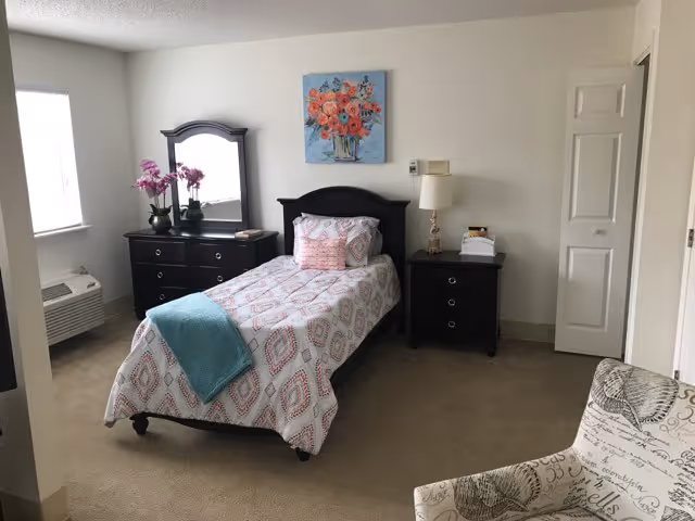 Bright furnished bedroom with a single bed, dark wood dresser and nightstand, upholstered chair, and floral wall art.