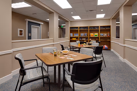 A common area in a senior living facility with several tables and chairs arranged for socializing or activities. One table has playing cards and snacks on it. In the background, there is a wooden bookshelf with books and board games. The room has beige walls, a carpeted floor, and recessed ceiling lights.
