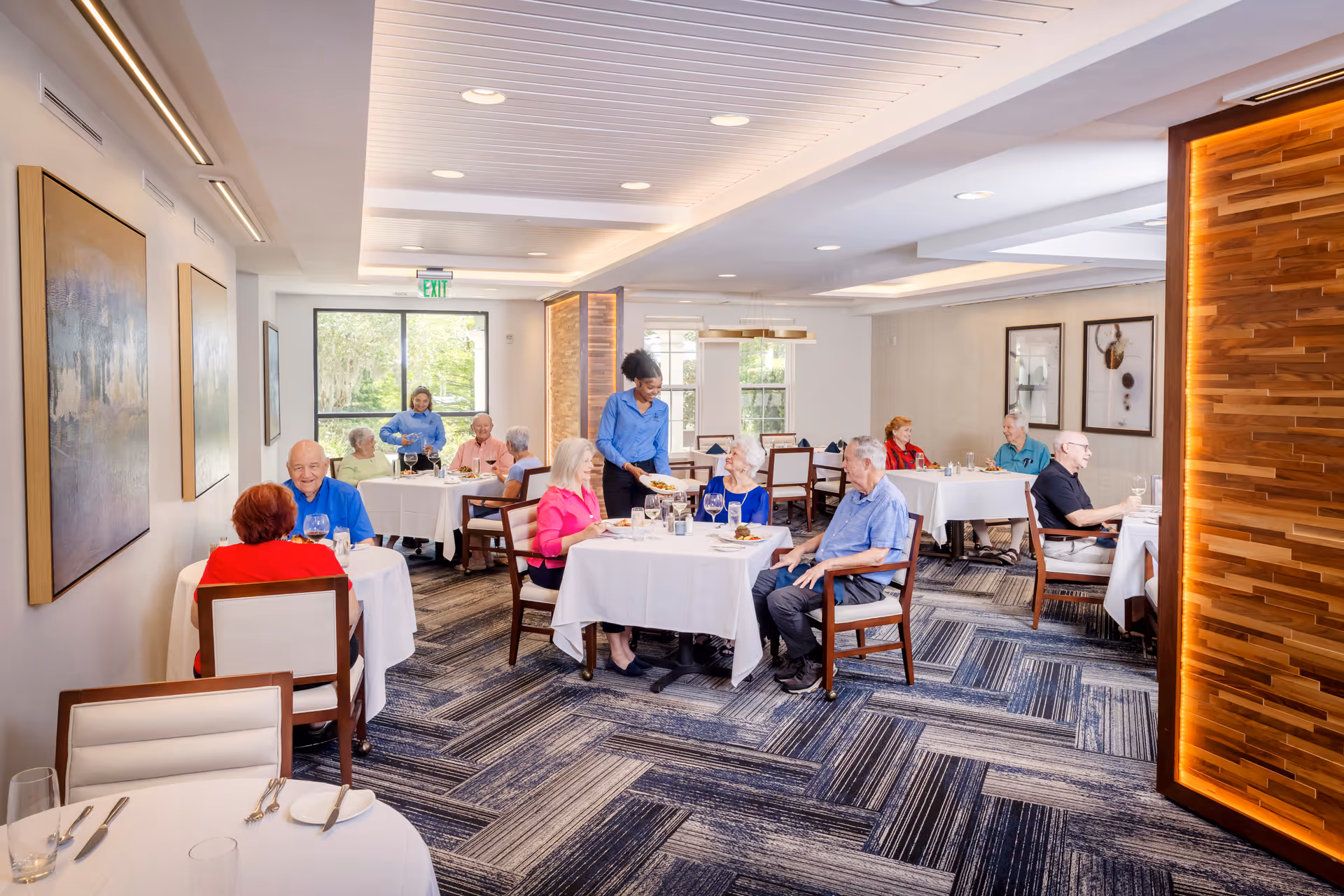 A bright and modern dining room with several elderly people seated at tables covered with white tablecloths. A server is attending to one of the tables. The room has large windows letting in natural light, contemporary artwork on the walls, and a patterned carpeted floor.