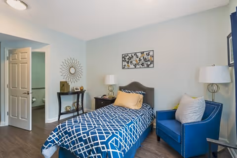 Furnished bedroom with a single bed, blue armchair, side tables, wall art and an open door to a bathroom.