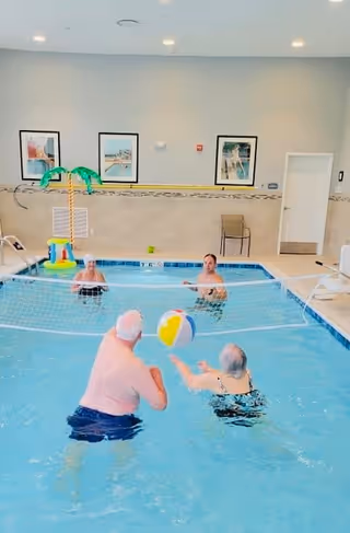 Four elderly people playing volleyball with a beach ball in an indoor swimming pool. The pool area has light-colored walls with framed pictures and a small inflatable palm tree decoration near the pool edge.