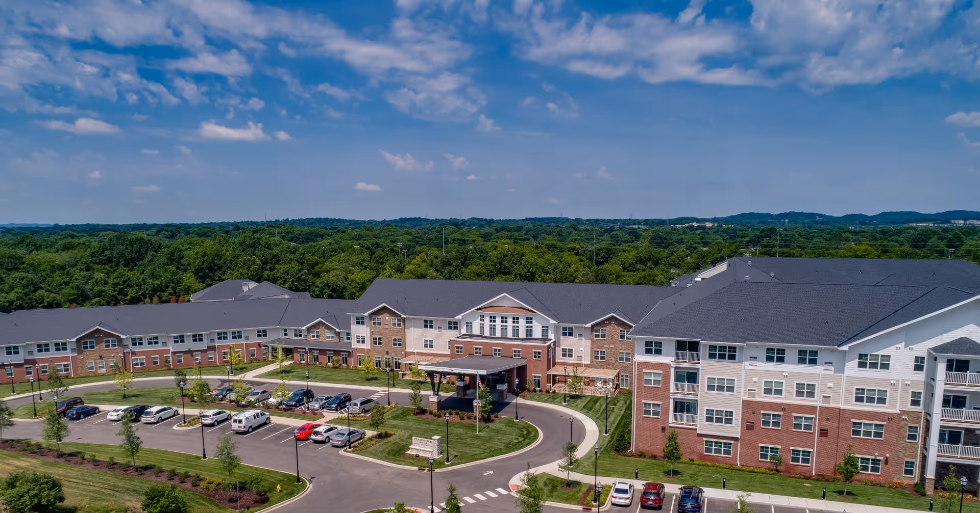 Aerial view of Clarendale at Indian Lake senior living facility showing a large multi-story building with brick and light-colored siding, surrounded by parking lots, landscaped greenery, and trees under a partly cloudy blue sky.