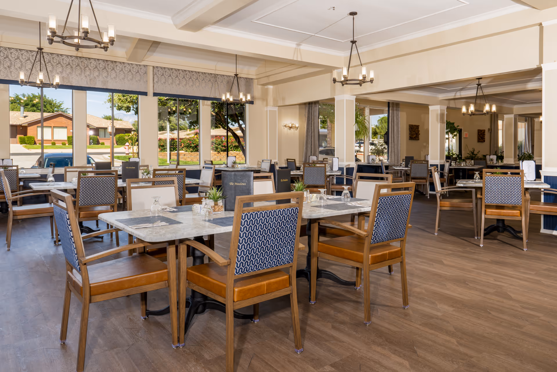 A spacious dining room with multiple tables and chairs arranged neatly. The tables have marble-like surfaces with place settings including glasses and napkins. Large windows allow natural light to fill the room, showing a view of trees and houses outside. The room features wooden flooring, chandeliers, and light-colored walls with decorative molding.