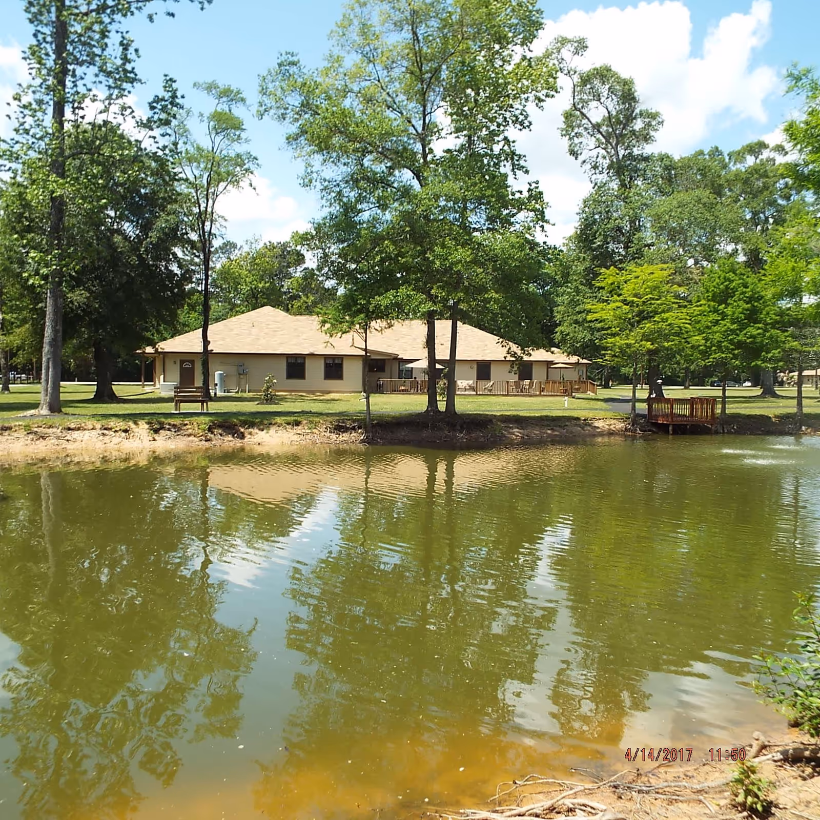 A single-story building with a beige roof and light-colored walls is situated behind a pond. The pond reflects the building and surrounding trees. There are several trees and a wooden bench near the water, with a small wooden dock extending into the pond. The sky is partly cloudy with blue patches.