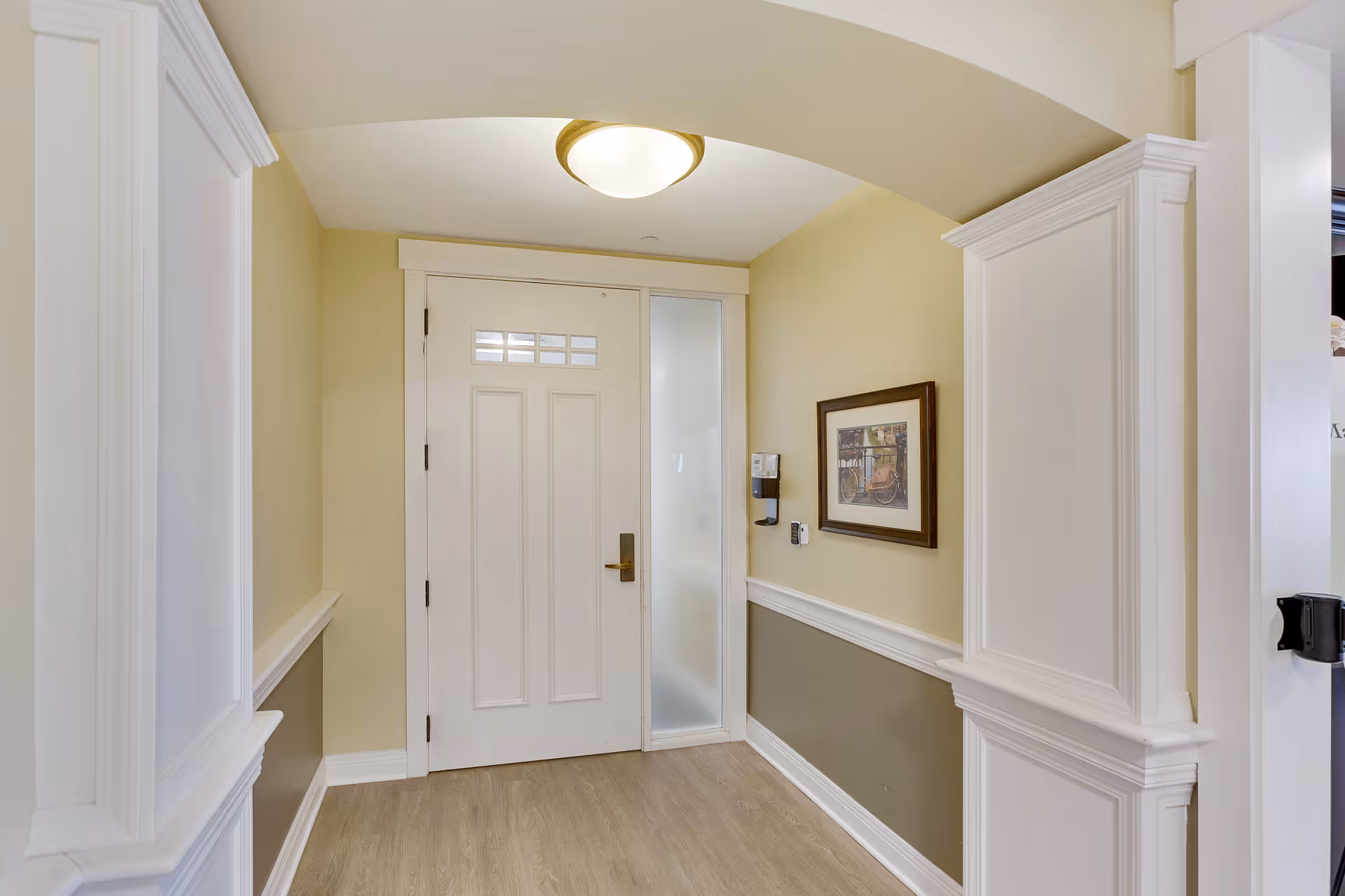 Interior view of a hallway with a white door at the end, beige walls with white trim, a framed picture on the right wall, and a ceiling light fixture.
