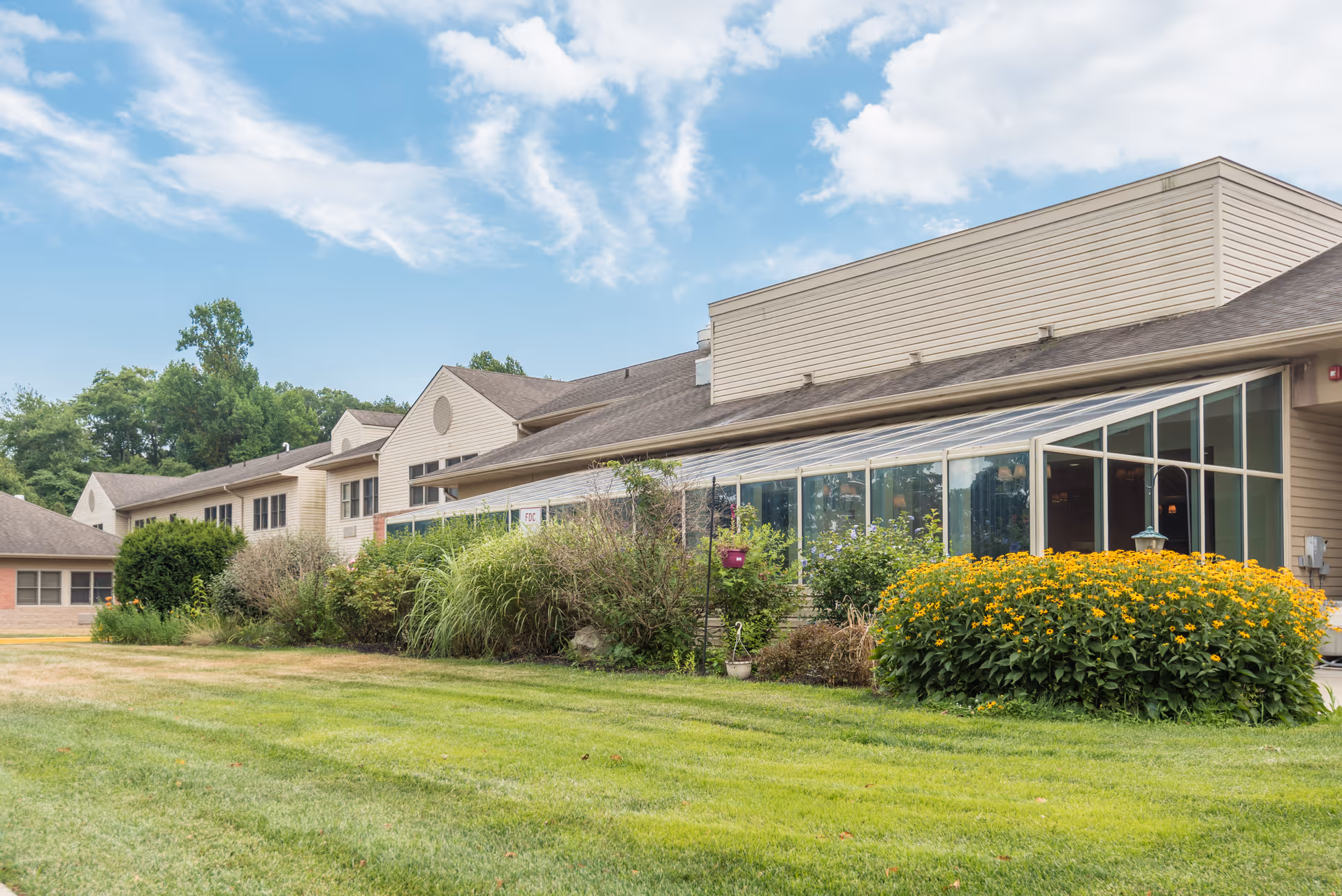 Exterior view of a senior living facility building with beige siding, large windows, and a well-maintained lawn with bushes and yellow flowers under a partly cloudy blue sky.