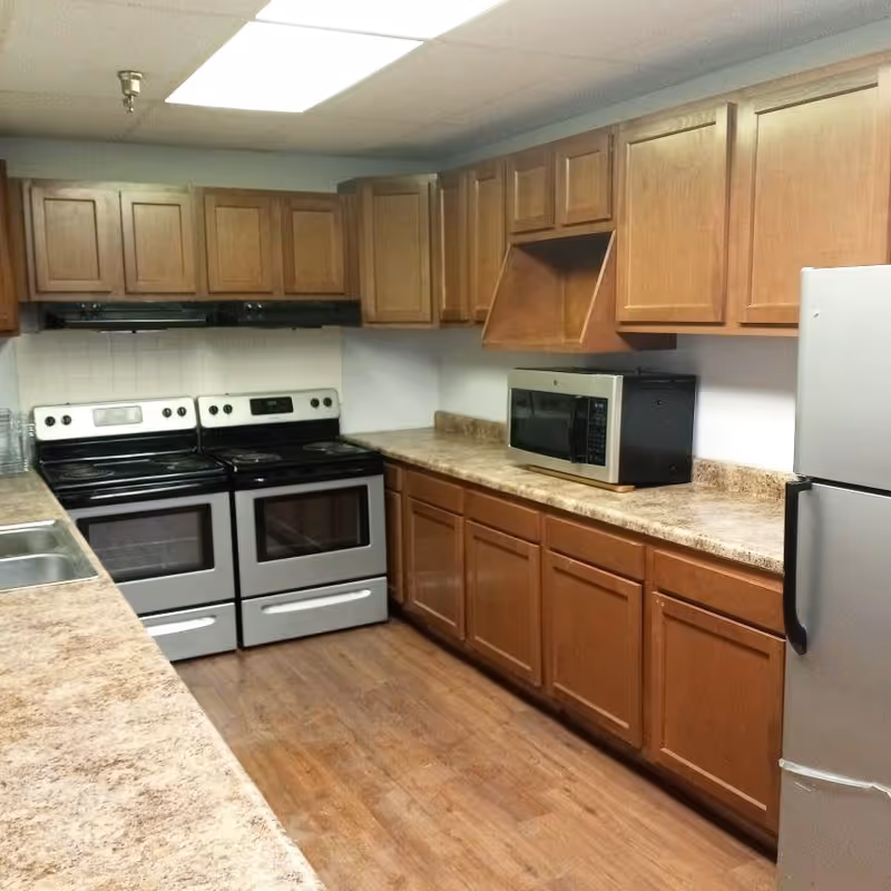 A kitchen with wooden cabinets, two electric stoves, a microwave on the countertop, a stainless steel refrigerator, and a double sink. The floor is wooden and the ceiling has a fluorescent light panel.