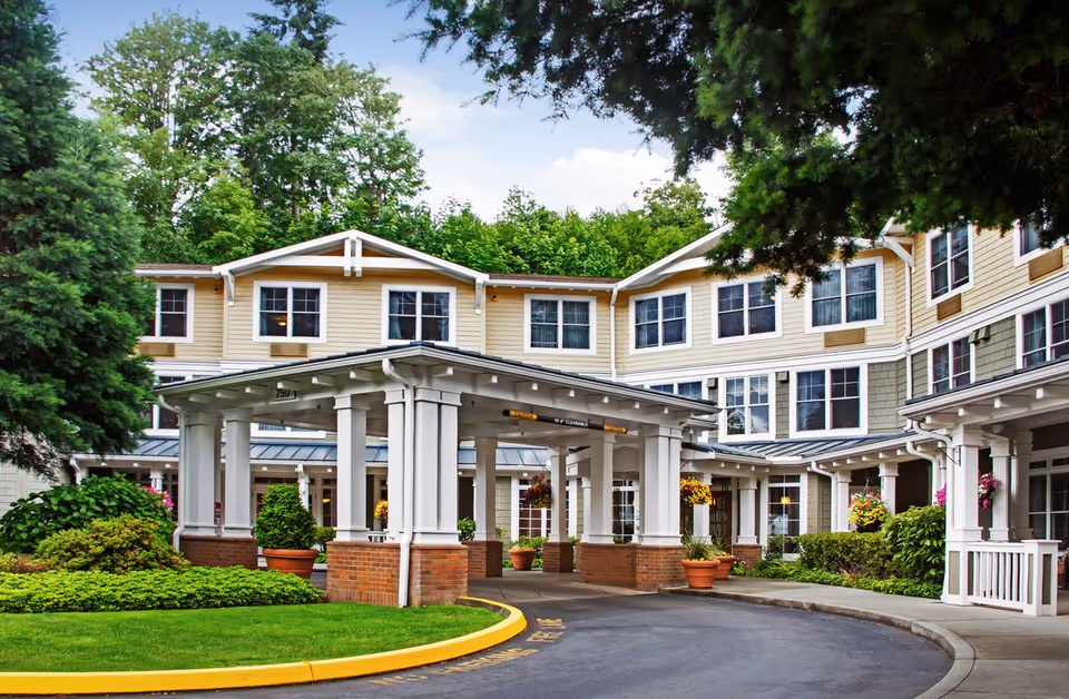 Exterior view of a senior living facility with a covered entrance, surrounded by greenery and potted plants, under a partly cloudy sky.
