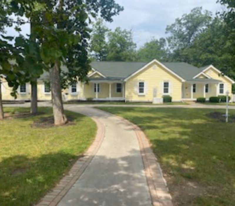 Front exterior of a single-story yellow senior living building with a curved walkway, lawn, and trees.
