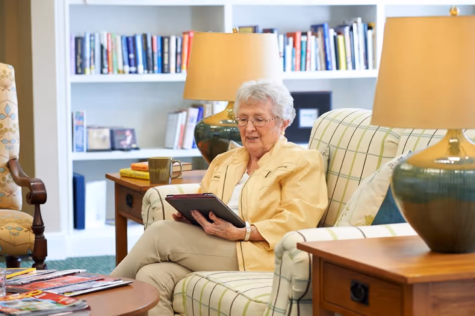 An elderly woman with white hair and glasses sits on a plaid sofa in a cozy living room, holding a tablet. The room features a wooden side table with a green lamp and a coffee mug, a bookshelf filled with books in the background, and a patterned armchair to the side.