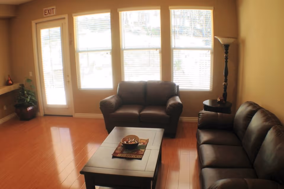 A cozy living room with two brown leather sofas facing each other, a wooden coffee table with a decorative bowl and cloth, three large windows with blinds, a tall floor lamp, a potted plant near a door with an exit sign above it, and wooden flooring.