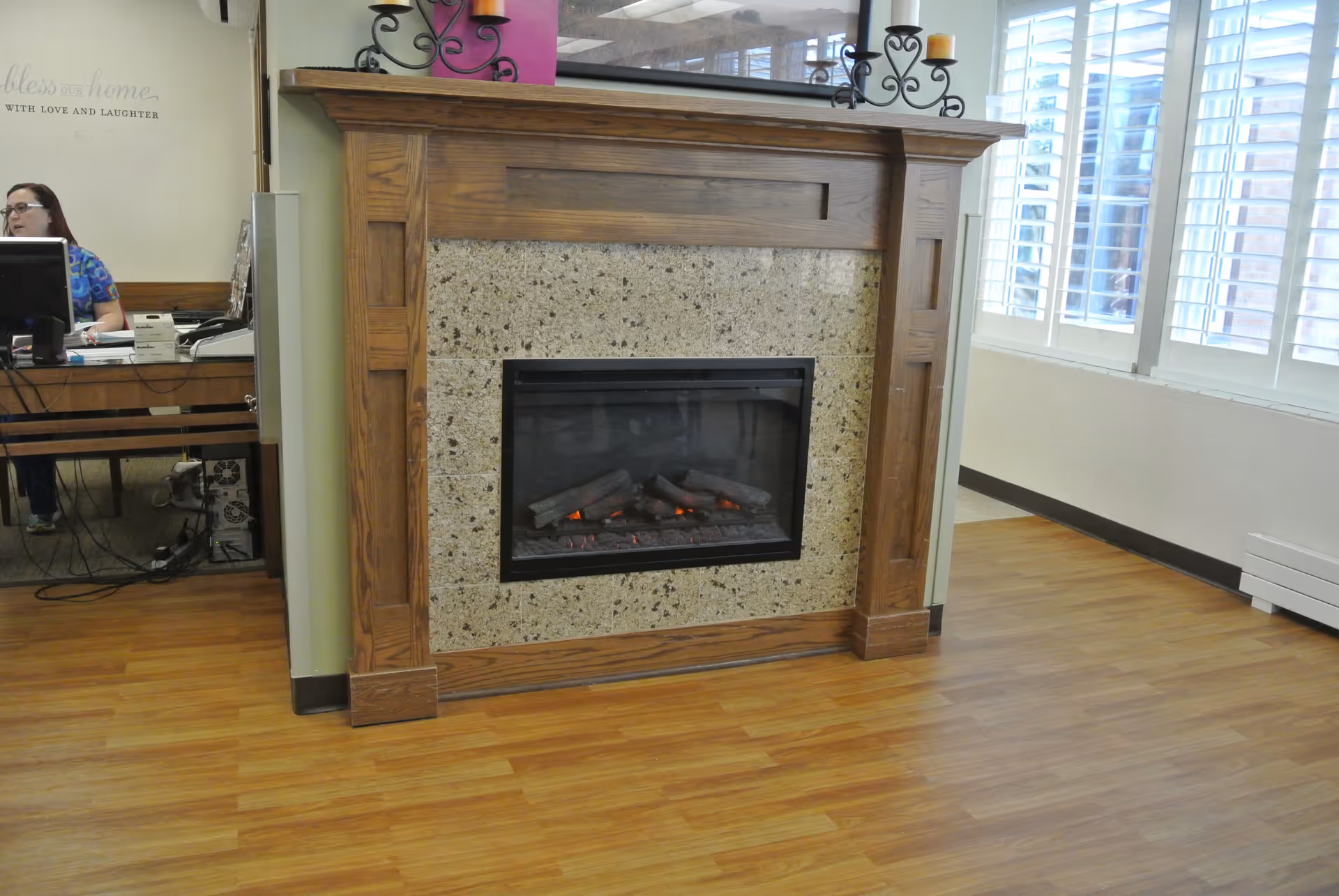 Interior view of a room with a wooden floor and a decorative fireplace with a stone surround and wooden mantel. To the left, a woman is seated at a desk with a computer. Large windows with white shutters are on the right side, allowing natural light into the room.
