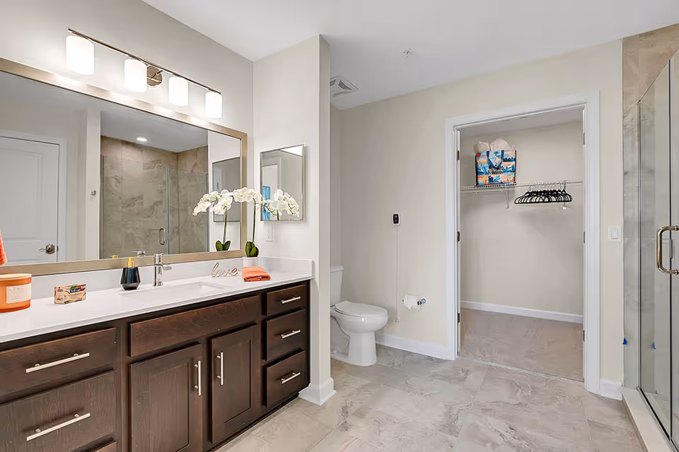 A modern bathroom featuring a large mirror above a dark wood vanity with a white countertop. On the countertop are decorative items including a candle, soap, a small plant with white flowers, and a towel. To the right is a white toilet and an open doorway leading to a walk-in closet with hangers and a bag on the shelf. The bathroom has a glass-enclosed shower with tiled walls and a tiled floor.