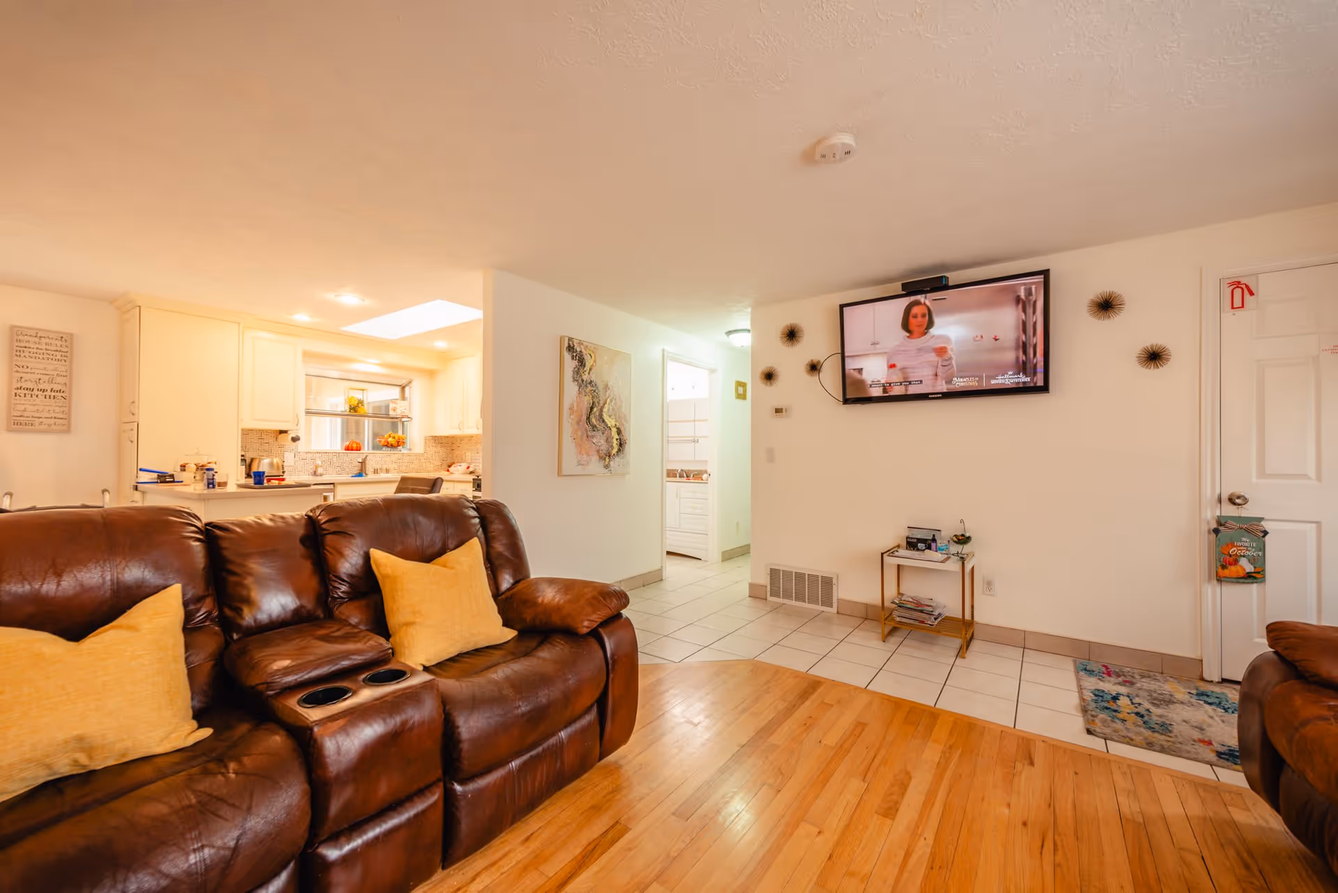 Interior view of a living room with brown leather sofas and yellow cushions, a wall-mounted TV showing a woman, a small table with magazines, and a kitchen area in the background with white cabinets and a tiled backsplash.