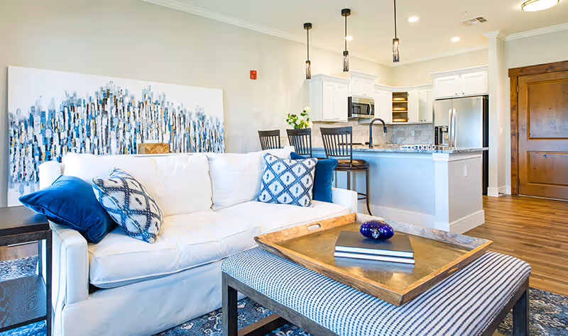 Bright and modern living room with a white couch adorned with blue and patterned pillows, a striped ottoman with a wooden tray holding books and a decorative item, and a kitchen area in the background featuring white cabinets, stainless steel appliances, and a breakfast bar with three chairs.