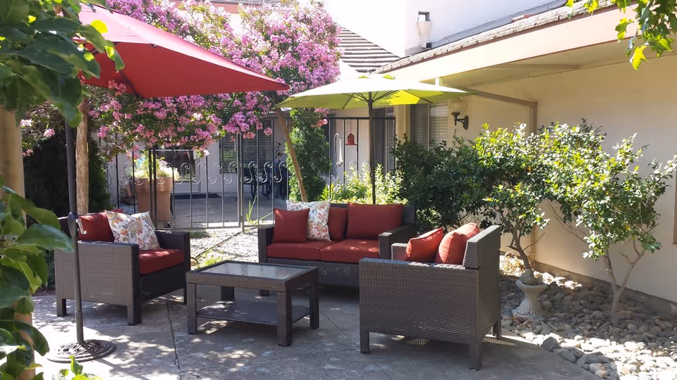 Outdoor patio area with wicker furniture including a sofa and two armchairs with red cushions and floral pillows. There is a glass-top coffee table in the center. Two large umbrellas, one red and one green, provide shade. The area is surrounded by greenery and flowering trees with pink blossoms, next to a building wall.