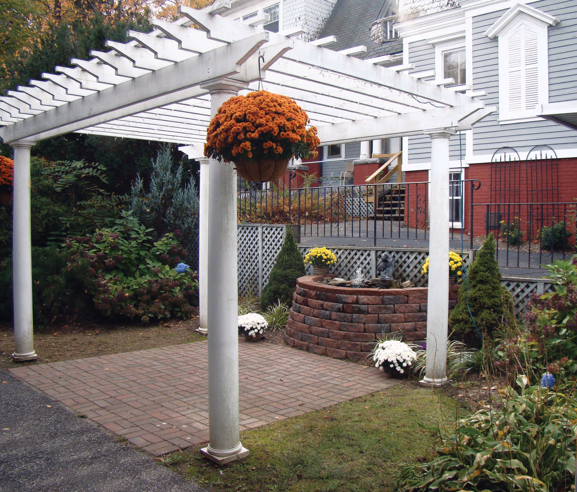 Outdoor garden area at Wolcott Hall Nursing Center featuring a white pergola with a hanging basket of orange flowers, a circular brick planter with yellow and white flowers, and various shrubs and plants surrounding the space. The background shows part of a gray and red building with stairs and a black metal railing.