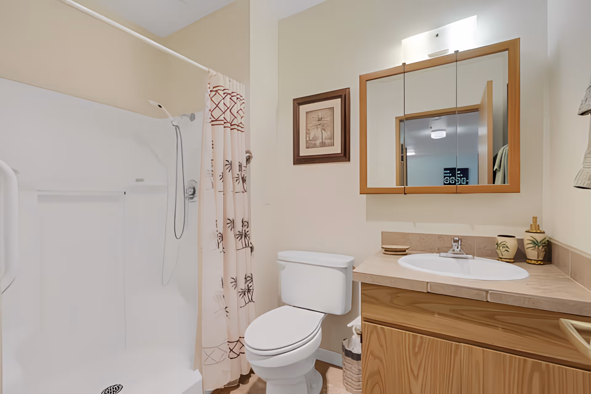 A clean bathroom featuring a white toilet, a wooden vanity with a sink, a mirror with wooden frame above the sink, a shower with a white curtain decorated with palm tree patterns, and a framed picture on the wall.