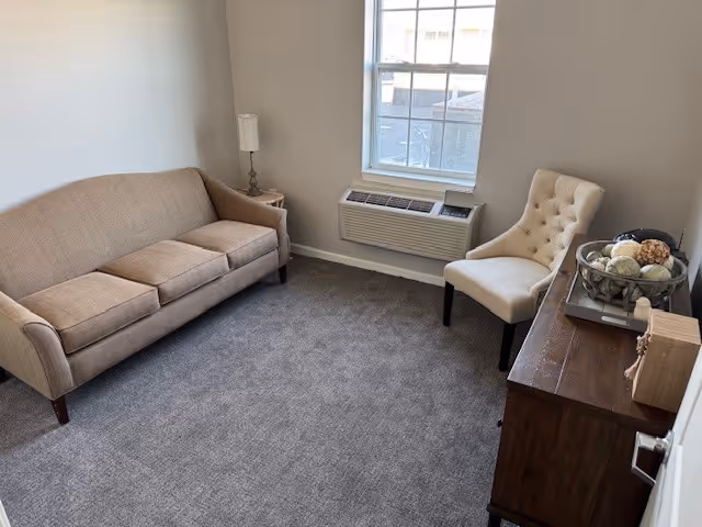 Small furnished living room with a beige sofa, tufted chair, window air conditioner, and wooden dresser with decorative bowl.