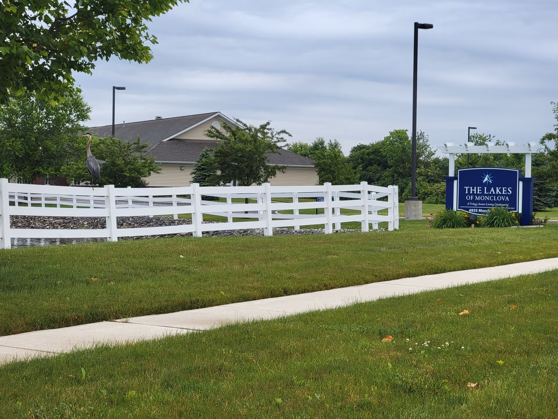 The entrance to The Lakes of Monclova with a white fence, grassy lawn, pond, and the facility sign.