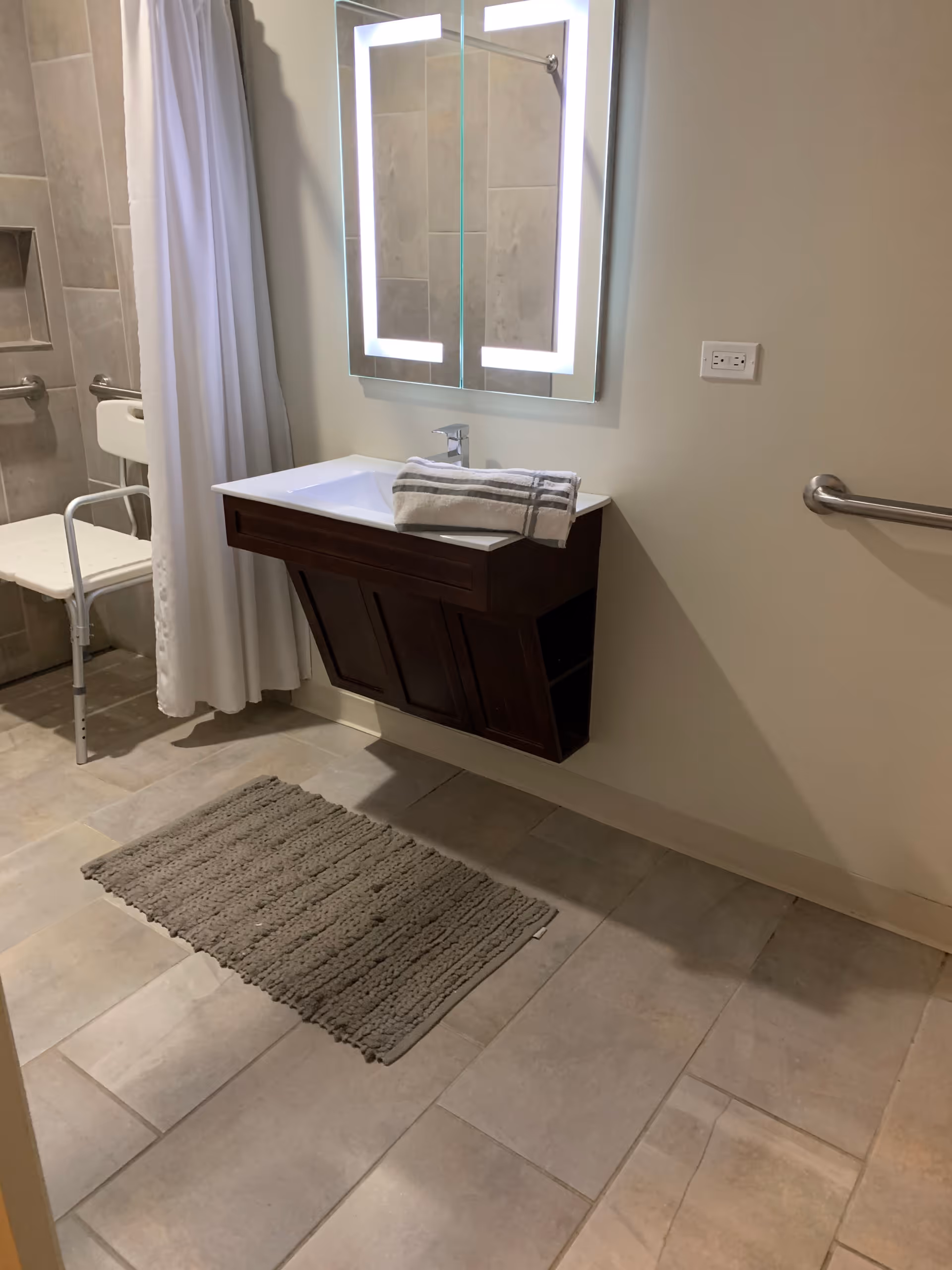 A modern bathroom with beige tiled floor and walls, featuring a wall-mounted dark wood vanity with a white sink and a folded towel on top. Above the sink is a rectangular mirror with built-in LED lighting. To the left, there is a white shower curtain and a white shower chair with metal legs. A gray textured bath mat is placed on the floor in front of the sink. A metal grab bar is mounted on the right wall.