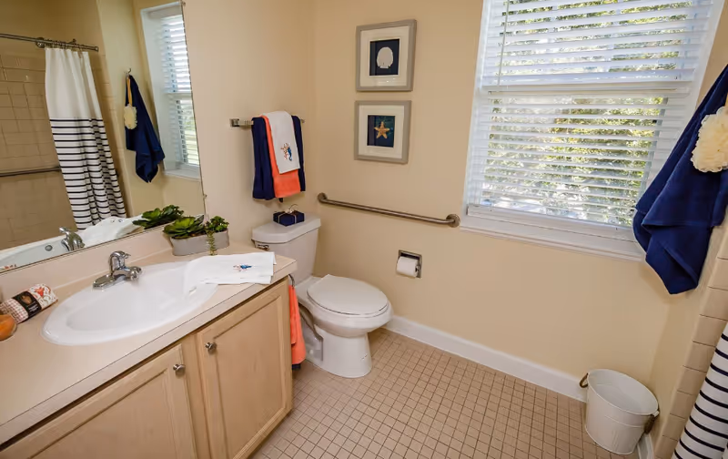 Bright bathroom with a sink and vanity, toilet with a safety grab bar, striped shower curtain, and a window with blinds.