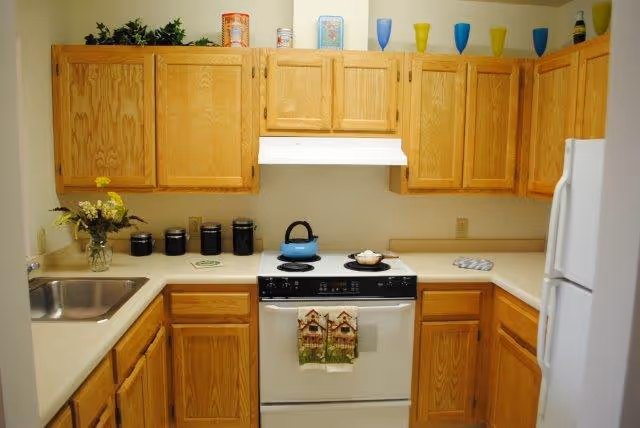 A compact kitchen with wooden cabinets, a white stove with a blue kettle on top, a white refrigerator, and a stainless steel sink. The countertop has a vase with flowers, several black canisters, and a few decorative items on top of the cabinets including colorful cups and small containers.