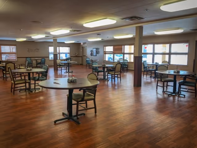 A spacious dining room with several round tables and chairs arranged neatly on a wooden floor. Large windows with blinds allow natural light to fill the room. The walls are decorated with a few pieces of artwork and a wall decal. The room appears clean and well-lit.