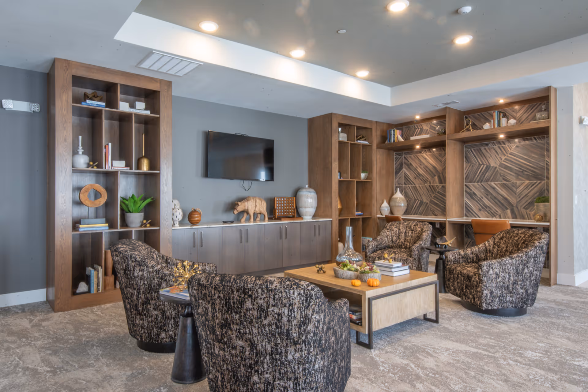 Contemporary lounge with four patterned armchairs around a wood coffee table, built-in shelving, and a wall-mounted TV.