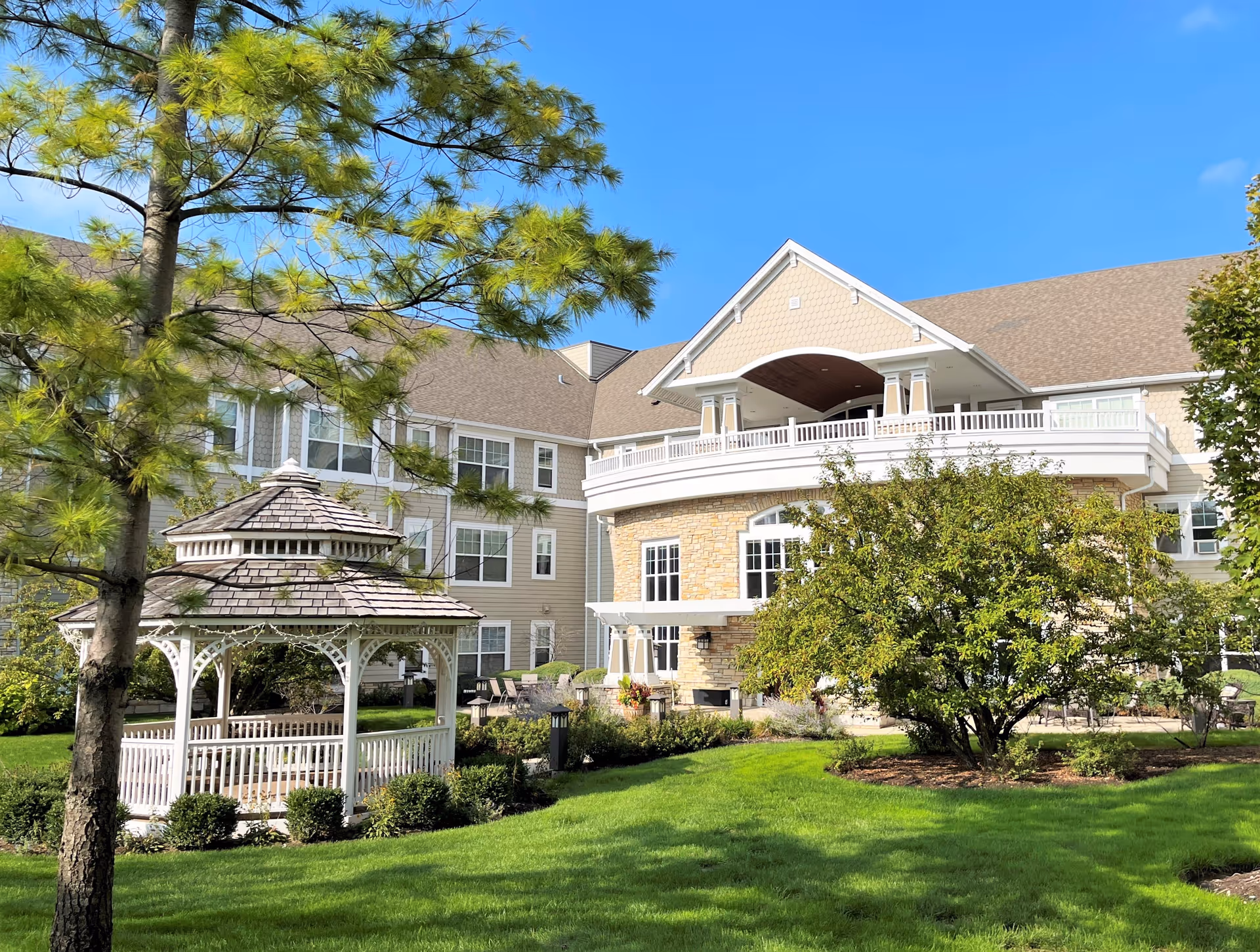 Outdoor view of Chestnut Square at The Glen featuring a well-maintained green lawn, a white wooden gazebo, trees, and a multi-story building with beige siding and stone accents under a clear blue sky.