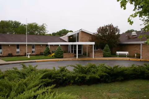 Single-story brick senior living facility entrance with a covered canopy and landscaped shrubs.