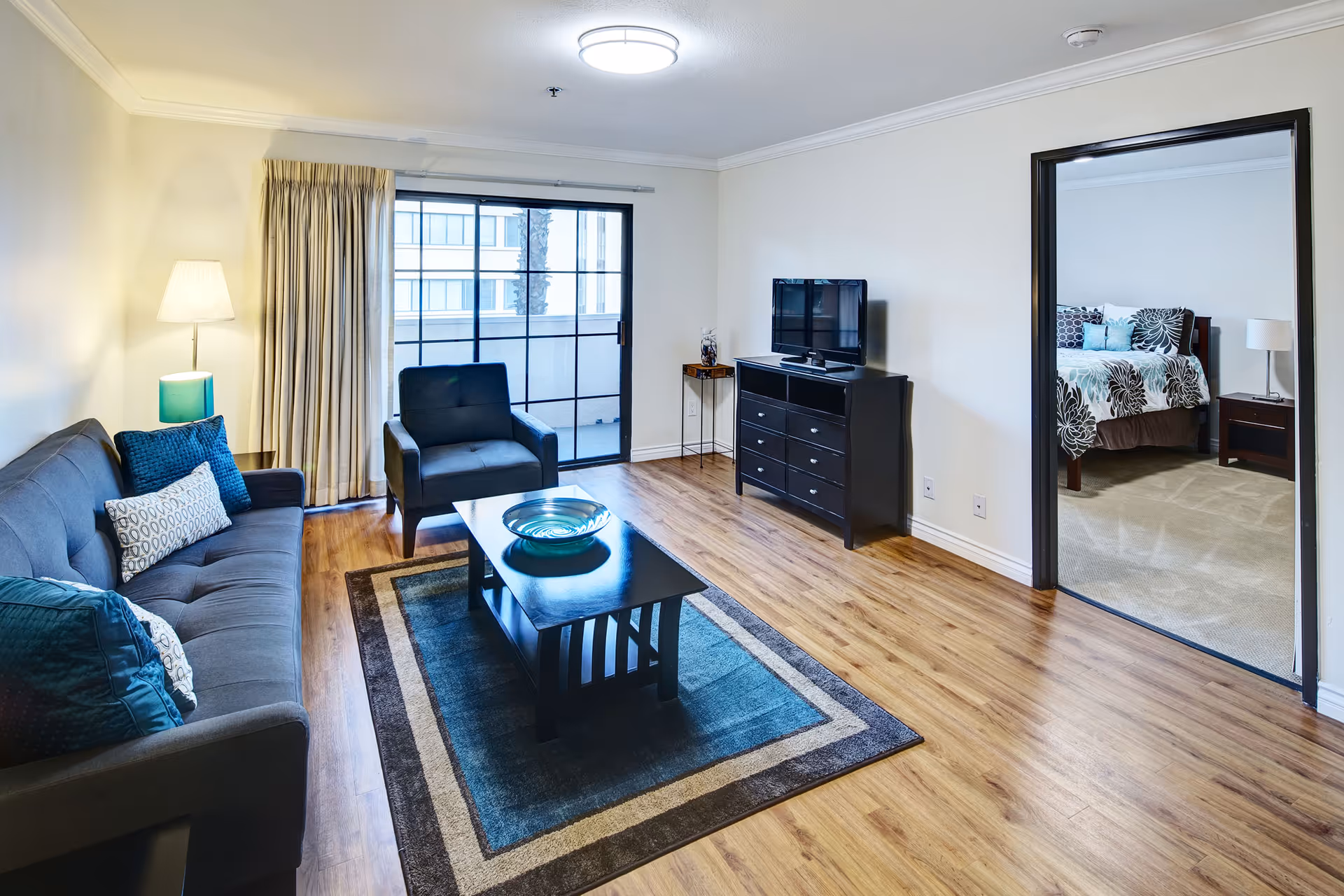 A bright living room with wooden flooring featuring a gray sofa with blue and patterned cushions, a matching armchair, a black coffee table with a decorative bowl, and a patterned area rug. A flat-screen TV sits on a black dresser against the wall. There is a sliding glass door with beige curtains leading to a balcony. An open doorway reveals a bedroom with a bed covered in floral bedding and a nightstand with a lamp.