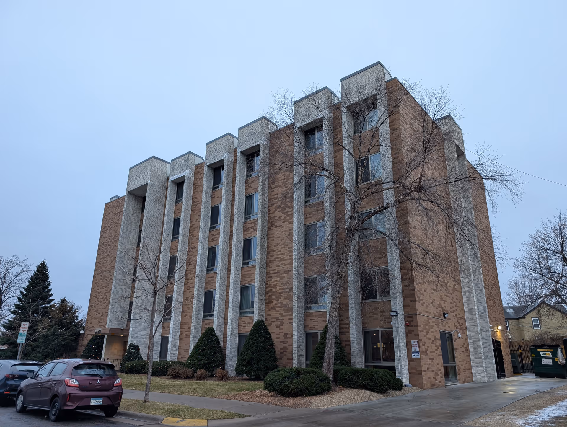 Exterior view of a multi-story brick building with white vertical accents, several windows, and some leafless trees and bushes around it. There are parked cars along the street in front of the building under a cloudy sky.