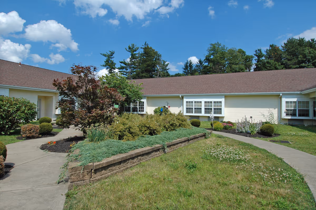 Outdoor courtyard area of a senior living facility with a paved walkway, landscaped garden beds, bushes, and trees. The building has light yellow siding and multiple windows under a brown shingled roof. The sky is blue with some clouds.