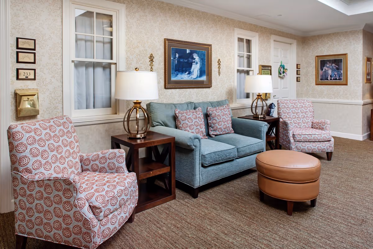 Cozy sitting area with two patterned armchairs, a blue loveseat, side tables with lamps, framed artwork, and a round leather ottoman.