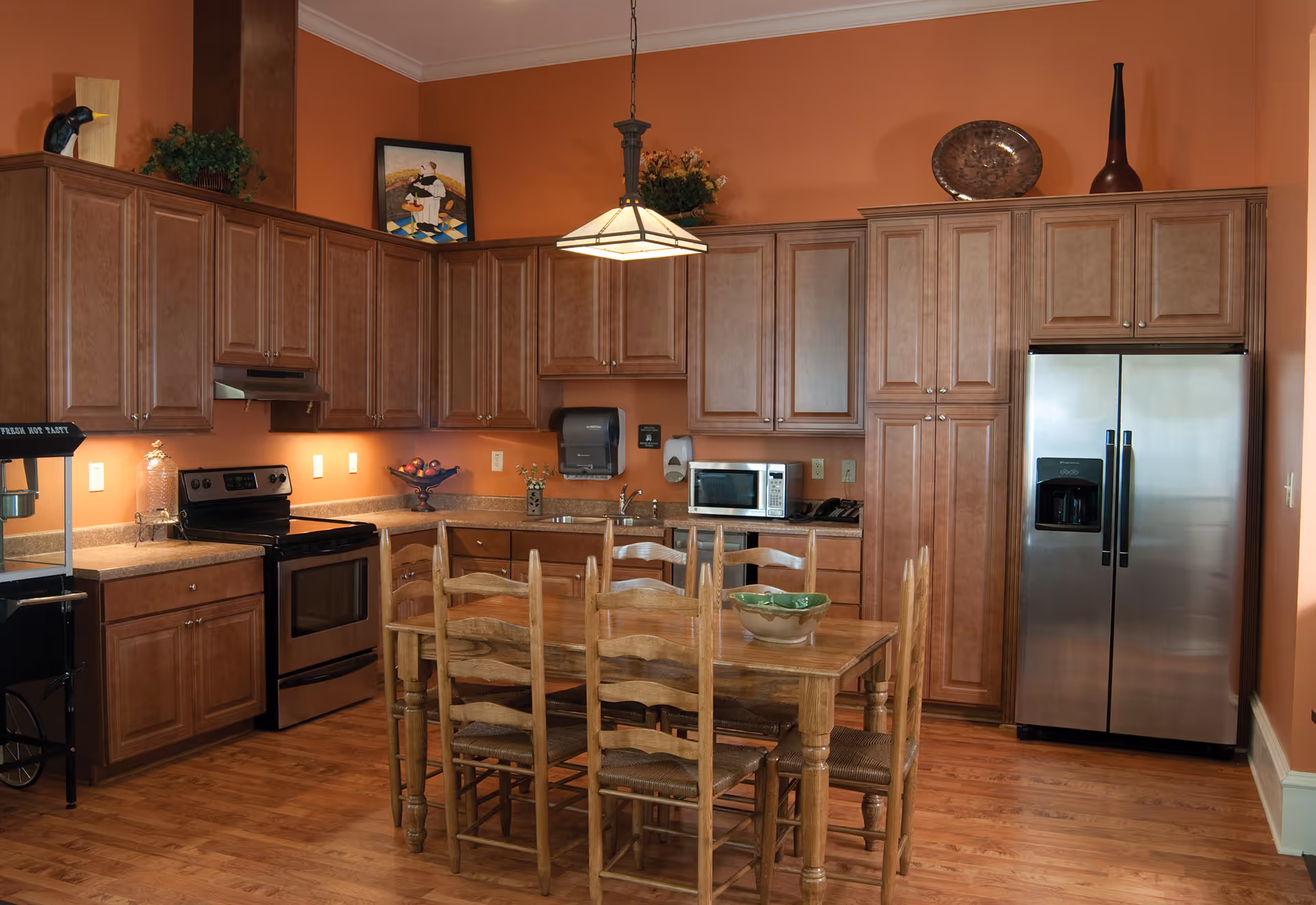 Warm-toned kitchen with wooden cabinets, stainless steel refrigerator and stove, and a wooden dining table with six chairs.