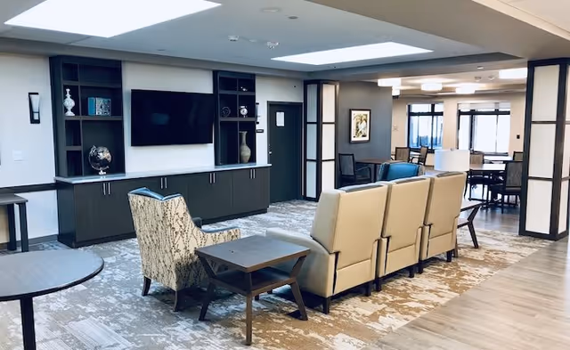 Spacious senior living communal lounge with recliner chairs, armchairs, side tables, a wall-mounted TV and built-in shelving, and a dining area in the background.