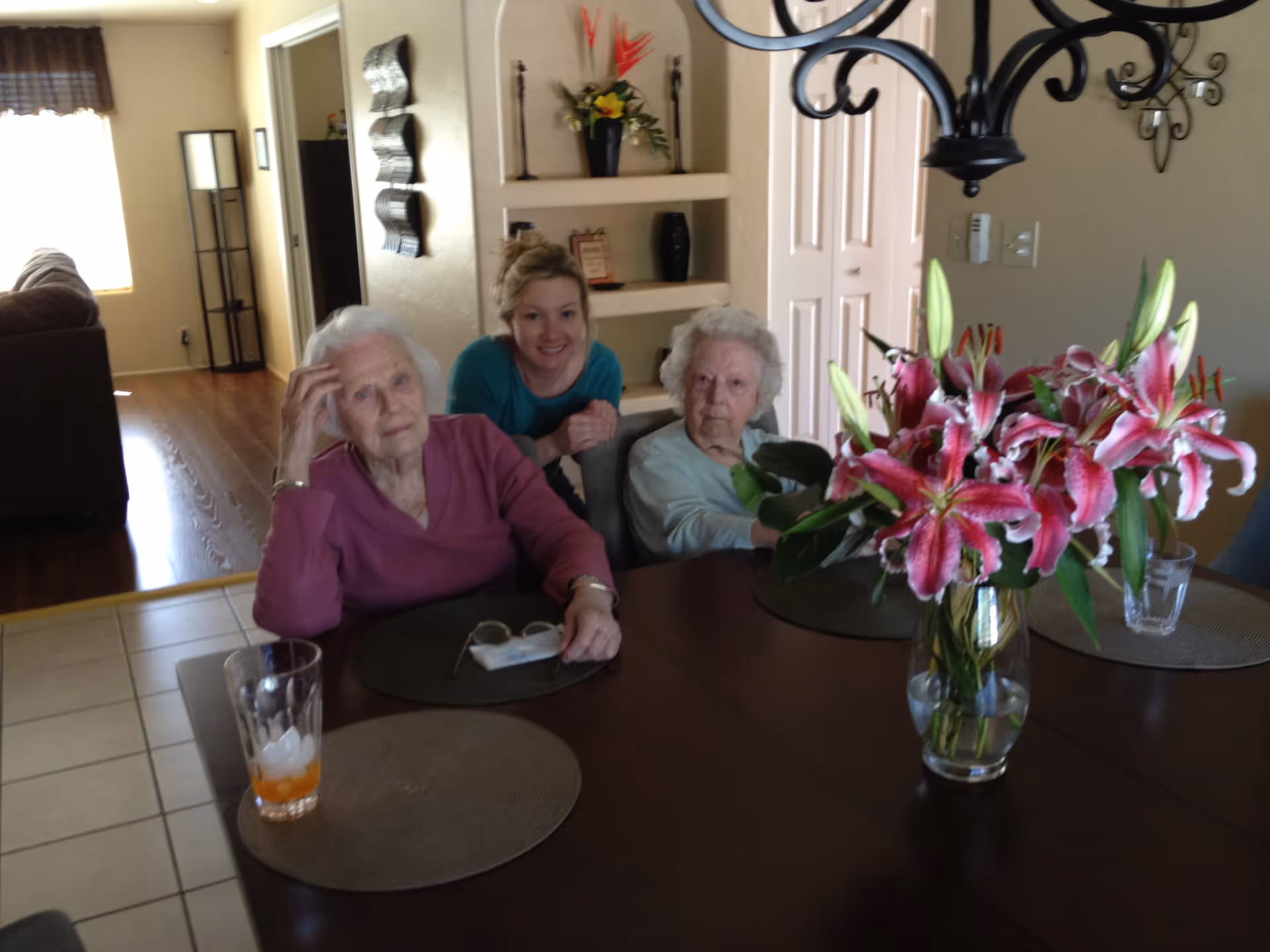 Three women — two elderly and one younger — seated at a dining table with a vase of pink lilies in a home dining room.