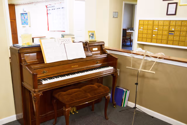 An upright wooden piano with sheet music on the stand and a cushioned bench in front. To the right, there is a music stand and some folders on the floor. Behind the piano, there is a wall with a calendar and a bulletin board. In the background, there is a wall with multiple mailboxes and a doorway leading to another room with a table and chairs.