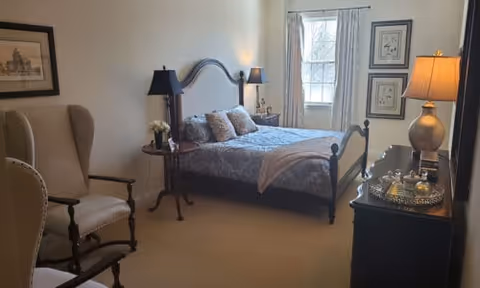 A cozy bedroom with a large bed featuring a decorative headboard, multiple pillows, and a throw blanket. There are two bedside tables with lamps on either side of the bed. A window with curtains allows natural light into the room. The room also contains a cushioned armchair, framed artwork on the walls, and a dresser with a lamp and decorative items on top.