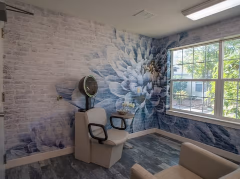 A small room with a large floral mural on the walls, featuring a blue and white flower design. The room contains two beige chairs, one with a vintage hair dryer hood attached, and a small table with a decorative item. A window on the right side lets in natural light and shows greenery outside.