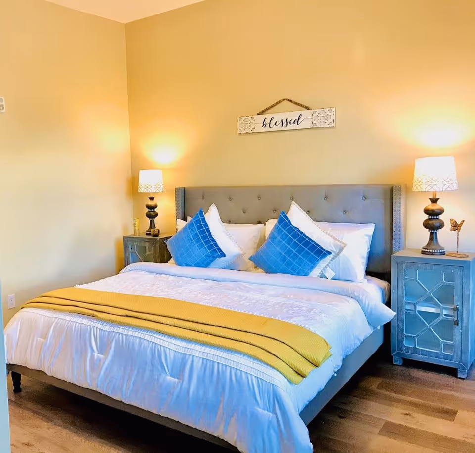 A neatly made bed with a gray upholstered headboard, white bedding, two blue decorative pillows, and a yellow throw blanket at the foot. On each side of the bed are matching bedside tables with lamps. Above the bed is a wall decoration with the word 'blessed'. The room has light yellow walls and wooden flooring.