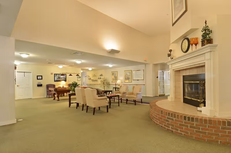 A spacious senior living facility common area with beige walls and carpet. The room features a large brick fireplace on the right, several upholstered chairs arranged around small tables, framed artwork on the walls, and a pool table in the background. Soft ceiling lights illuminate the space.