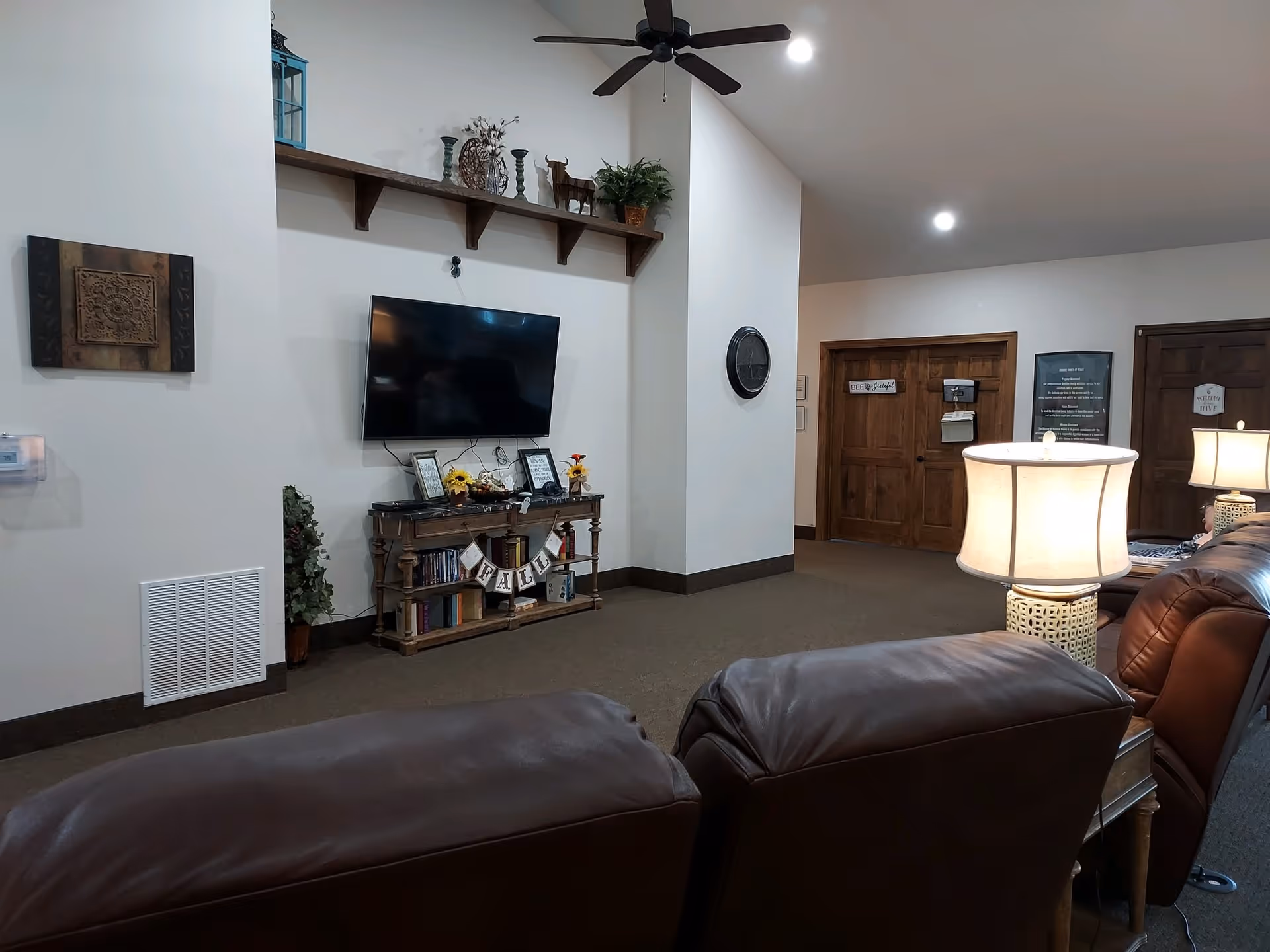 Interior view of a living room area with brown leather chairs, a wooden TV stand with decorative items and books, a wall-mounted flat screen TV, a ceiling fan, and wooden doors in the background. The room is softly lit with table lamps and has neutral-colored walls and carpet.